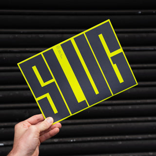 A hand holds a rectangular black sign with bold neon yellow lettering that reads 'SLUG' against a dark black corrugated metal background.