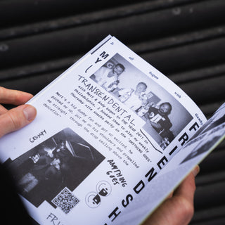 A person's hands holding an open printed magazine or booklet against a dark corrugated metal background. The page contains black-and-white photos, text, and a QR code.